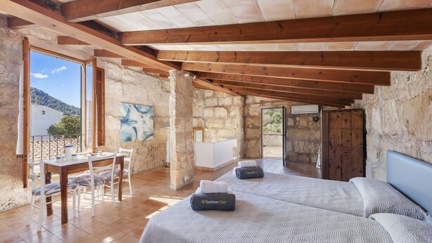 A rustic room with exposed stone walls, wooden beams, a dining area with a table and chairs, and two beds with towels. - Es Garroverar Villa: Luxurious Retreat in the Heart of Majorca (Pollensa)