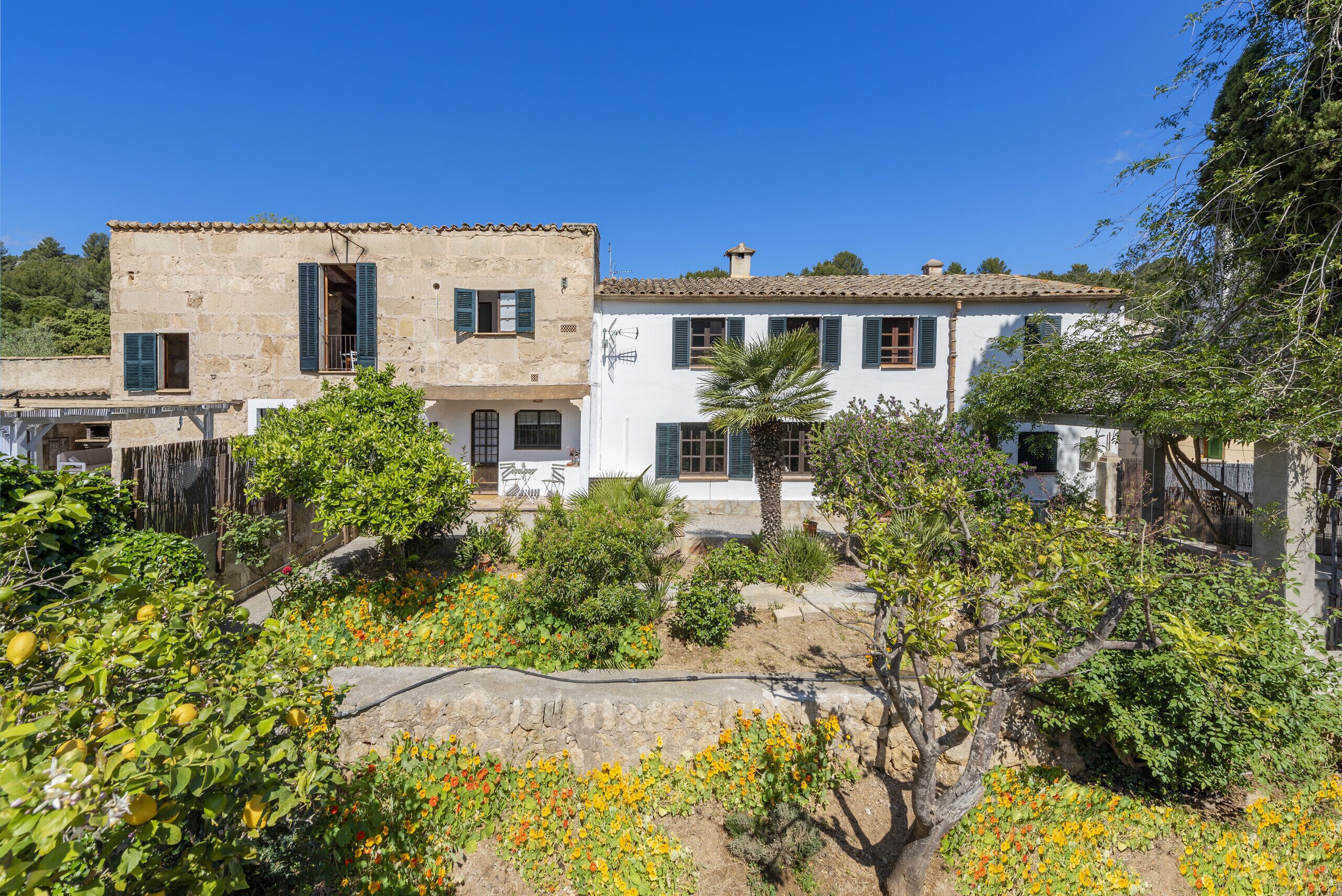 A two-story building with a stone facade and white walls, surrounded by lush greenery and flowering plants.