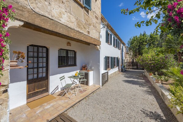 A narrow courtyard with a gravel path, a small outdoor seating area with a table and chairs, and a white building with shutters. - Es Garroverar Villa: Luxurious Retreat in the Heart of Majorca (Pollensa)