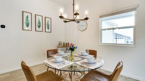 Un comedor con una mesa redonda de vidrio, sillas de cuero marrón, un candelabro, obra de arte con hojas enmarcadas, un reloj y una ventana con persianas.