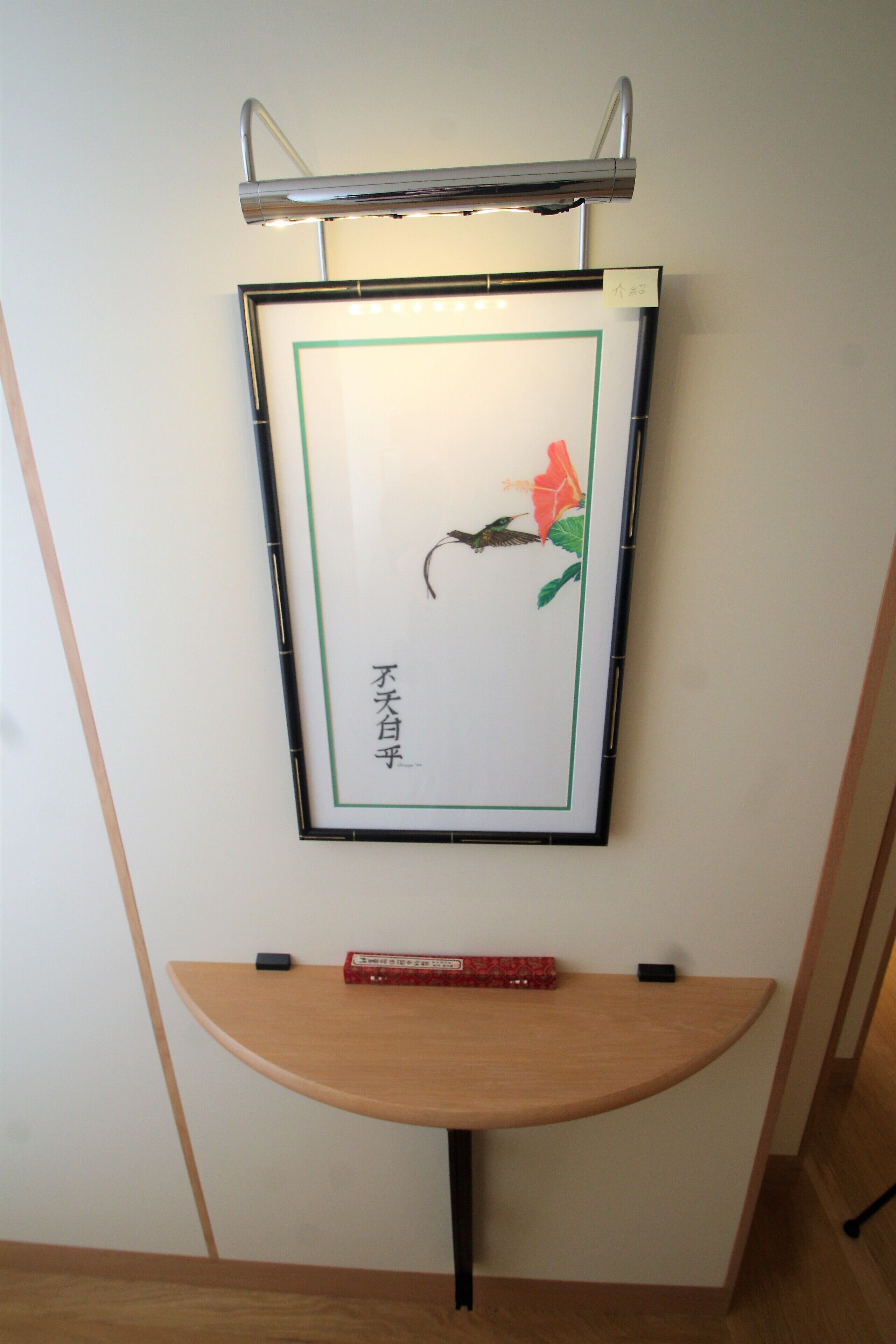 A framed artwork with a bird and flower on a wall, a small table below, and a light fixture above.