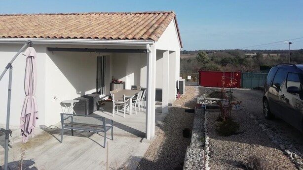 A white building with a tiled roof, a covered patio with outdoor furniture, and a gravel driveway. - Cosy home (Bassillac et Auberoche)