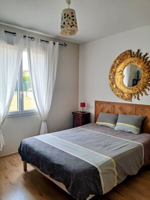 A bedroom with a bed, curtains, a nightstand, a lamp, a mirror with a sun design, and a window with a view.