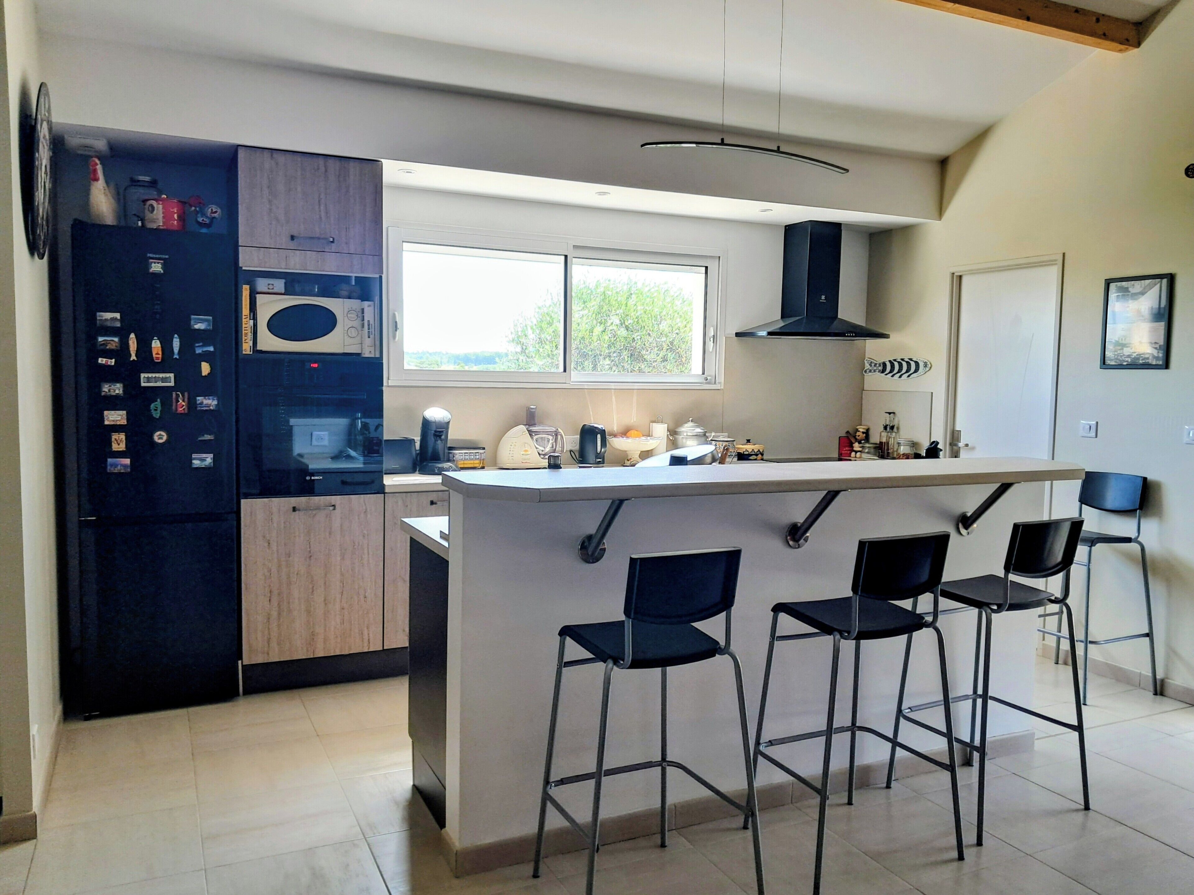 Une cuisine moderne dotée d’un îlot central, de tabourets de bar, d’un four encastré et d’une fenêtre laissant entrer la lumière naturelle.