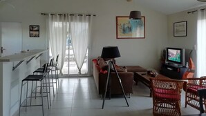 A spacious living area with a kitchen counter, bar stools, a sofa, a dining table with chairs, a tripod lamp, and a television. - Cosy home (Bassillac et Auberoche)