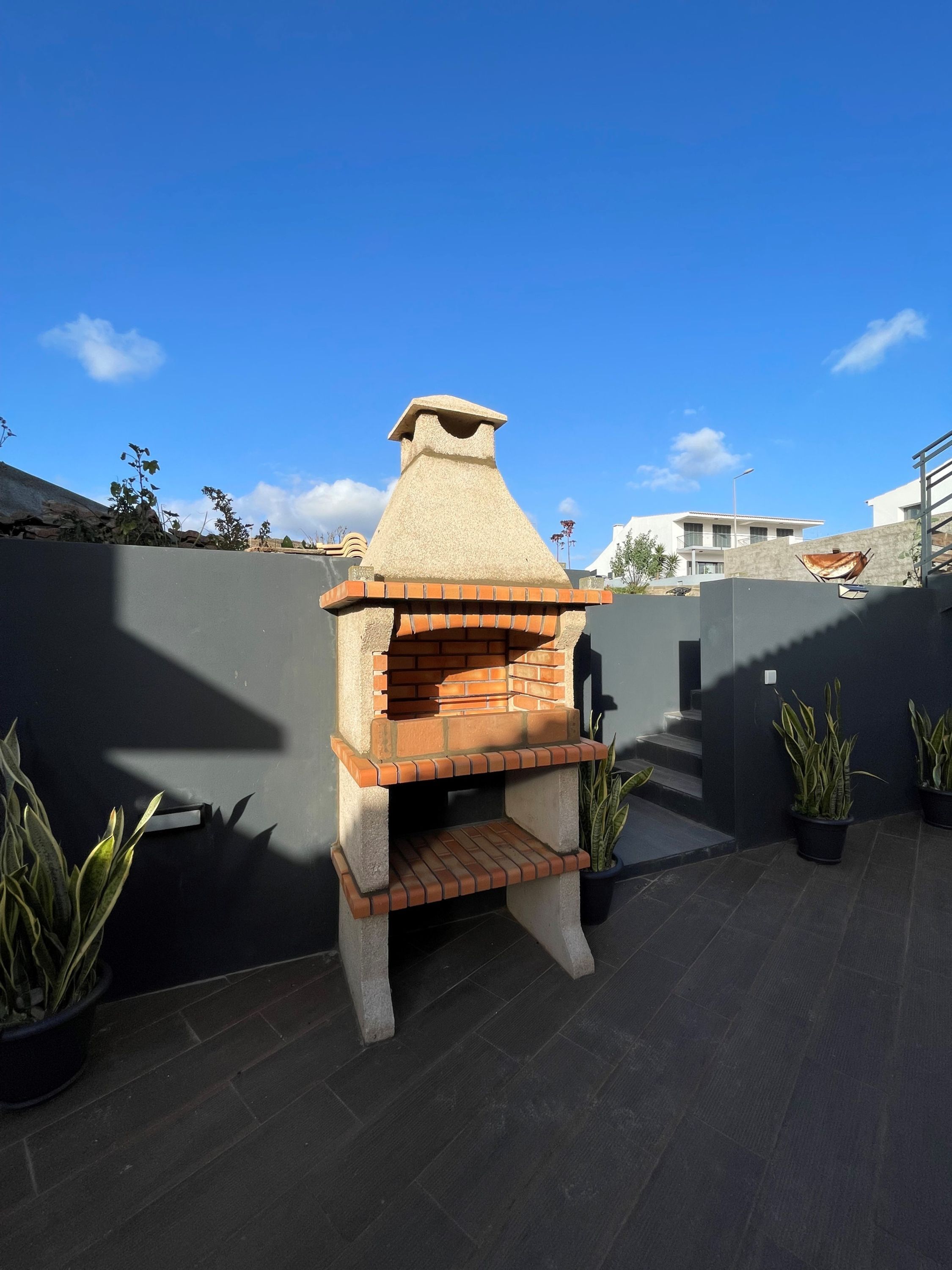 A brick-built outdoor oven with a chimney, surrounded by a dark wall and potted plants.