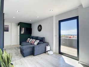 A modern living room with a grey sectional sofa, a mounted TV, a clock, and a view of the city through a large sliding glass door. - Casa da Figueira by Atlantic Holiday (Porto Santo)