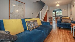 A living room with a blue sofa, yellow cushions, a map on the wall, a staircase, and a view to the outside. - Bumbles (Padstow)