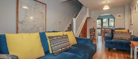 A living room with a blue sofa, yellow cushions, a map on the wall, a staircase, and a view to the outside.