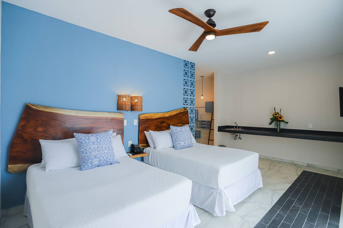 A hotel room with two beds, a ceiling fan, a nightstand with a lamp, and a small bathroom area with a sink and mirror.
