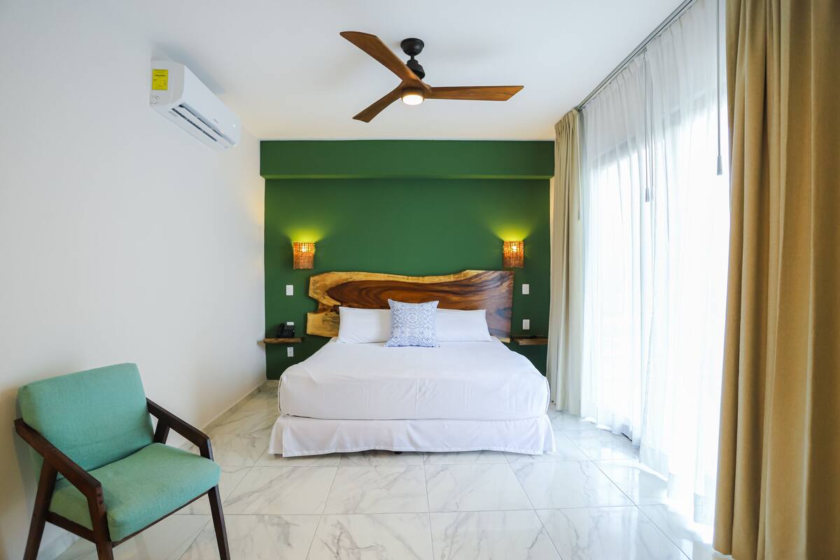 A hotel room with a bed, a chair, a ceiling fan, and a large window with curtains.