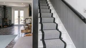 A staircase with grey carpeted steps and black handrails, leading to a landing with a fireplace and a dining area. - Ben View (Arrochar)
