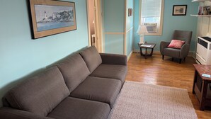 A living room with a brown sofa, a small table, a chair, a lamp, and a window with blinds. - Beachside Apt 1 (Frankfort)