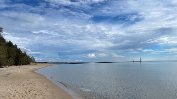 A beach with clear water, a lighthouse in the distance, and a forested area on the left. - Beachside Apt 1 (Frankfort)