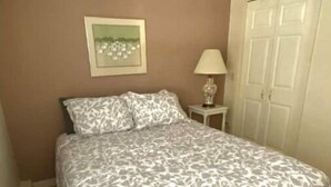 A bedroom with a bed, a nightstand, a lamp, a door, and a framed picture on the wall. - Beachside Apt 1 (Frankfort)