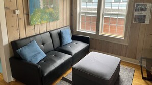 A room with a black leather sofa, a grey ottoman, wooden paneled walls, a large window with blinds, and a painting on the wall. - Beachside Apt 2 (Frankfort)