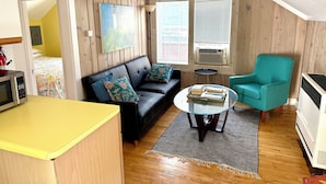 A room with a black leather sofa, a teal armchair, a small round table with books, a ceiling fan, and wooden walls. - Beachside Apt 2 (Frankfort)