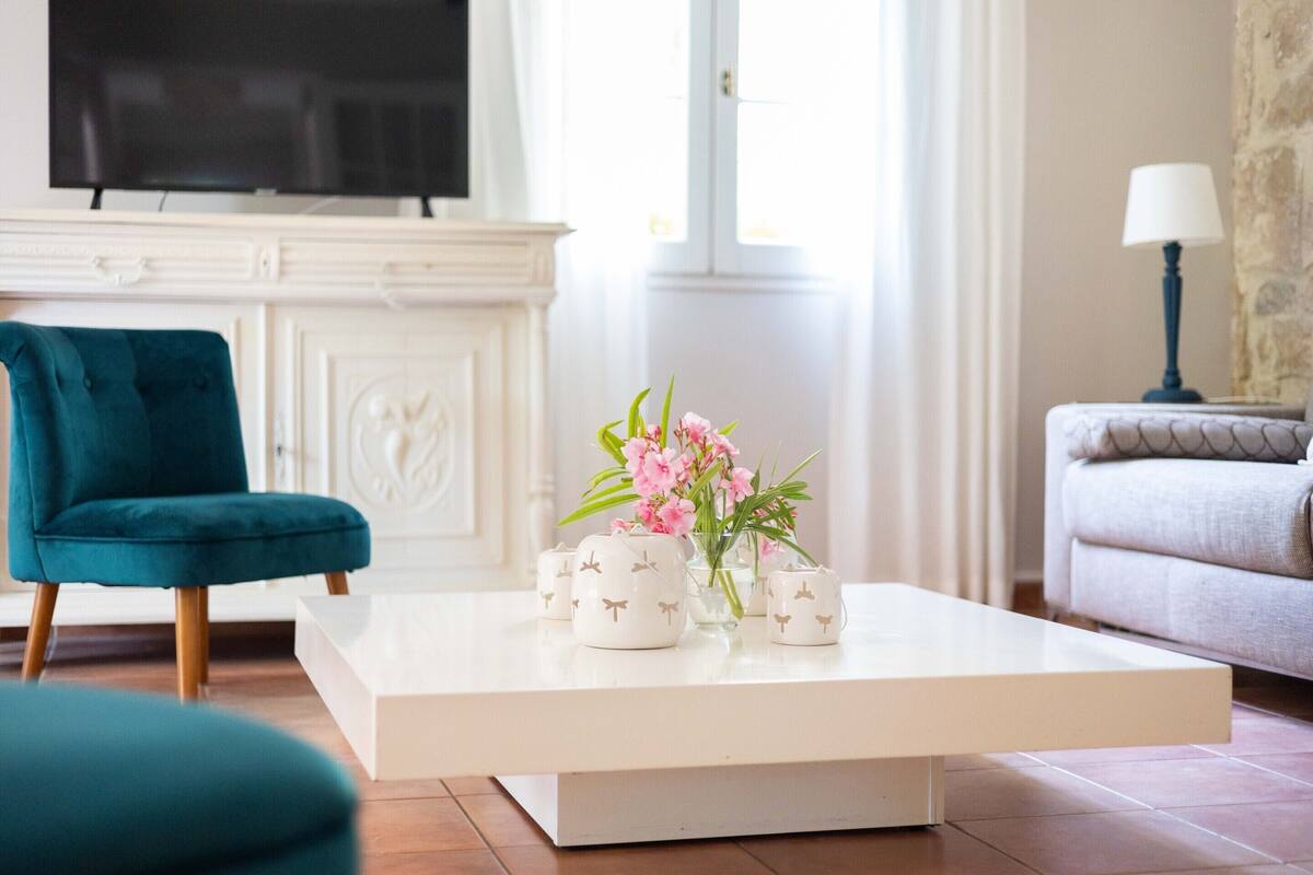 A living room with a teal chair, a white coffee table with floral vases, a television on a white mantelpiece, and a lamp on a side table.