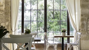 Ein rustikaler Raum mit Steinwänden, einem großen Fenster mit Vorhängen, weißen Stühlen und einem Holztisch.
