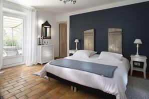 Un dormitorio con una cama grande, ropa de cama blanca, una pared de acento oscura, un cabecero de madera, mesitas de noche con lámparas, un espejo y vistas al exterior a través de una ventana con cortinas blancas.