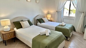 Ein Hotelzimmer mit zwei Einzelbetten, beide mit grüner Bettdecke und weißen Laken. An jedem Bett befindet sich ein Nachttisch mit einer Lampe. Das Zimmer hat ein Fenster mit weißen Vorhängen und die Wände sind in einem hellen Farbton gestrichen.