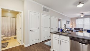A modern kitchen with white cabinets, a stainless steel dishwasher, and a built-in oven. There is a dining area with a dark countertop and a window with blinds.