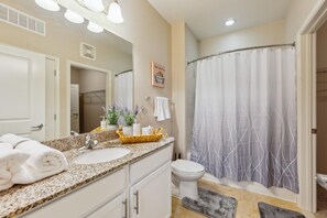 Une salle de bain avec un lavabo sur un comptoir en granit, des toilettes blanches, une douche avec un rideau à motifs et un miroir avec des lumières au-dessus.