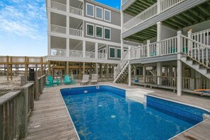 Una terraza de madera de varios niveles con una piscina, mobiliario exterior y una escalera que lleva a un balcón.