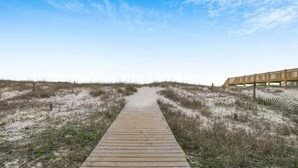 Un paseo marítimo de madera que conduce a una estructura de madera con barandilla, rodeada de dunas de arena y vegetación escasa.