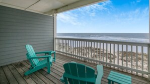 Una terraza techada con sillas Adirondack verdes y una pequeña mesa, con vista a una playa de arena y el océano.