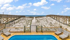 A wooden deck with a pool, overlooking a beach with sand dunes and a clear sky.