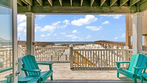 A wooden deck with two green Adirondack chairs, a white railing, and a view of the beach and ocean.