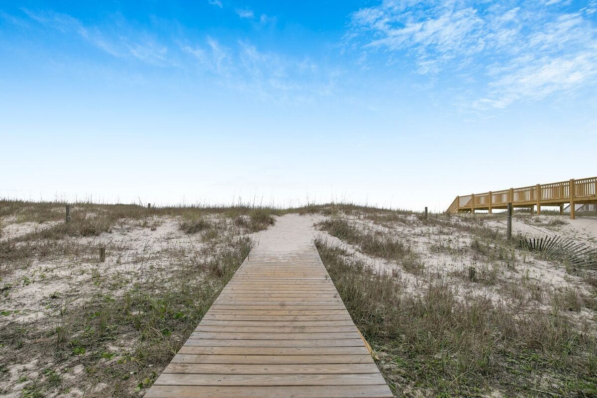 Een houten looppad dat leidt naar een houten gebouw met een reling, omringd door duinen en schaarse begroeiing.