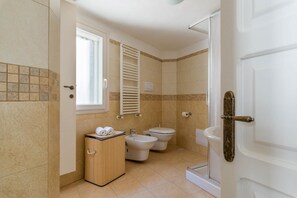 A bathroom with a toilet, bidet, and a sink. There is a cabinet with towels and a window.
