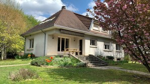Una casa de dues plantes amb un teulat inclinat, un porxo amb una banca i un petit jardí amb flors i arbustos.