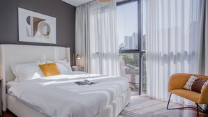 Une chambre d’hôtel moderne avec un grand lit, une table de chevet, une lampe de lecture, une chaise et une fenêtre avec des rideaux.