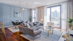Un salon moderne avec un canapé gris, un plancher en bois, un coin repas comprenant une table et des chaises, et une grande fenêtre donnant sur la ville.