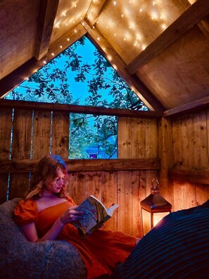 A cozy wooden cabin with a sloped ceiling, a window with a view of trees, and a person reading a book.