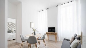 Een moderne woonkamer met een grijze bank, een televisie op een houten dressoir, en een eethoek met een ronde tafel en grijze stoelen.