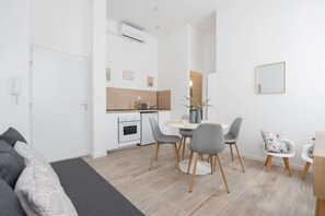 A modern living room with a sofa, a dining table with chairs, a kitchen area with a stove, and a large mirror on the wall.