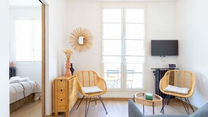 Une pièce moderne et bien éclairée, comprenant un lit, deux fauteuils en osier, un petit bureau en bois, une télévision et une grande fenêtre.
