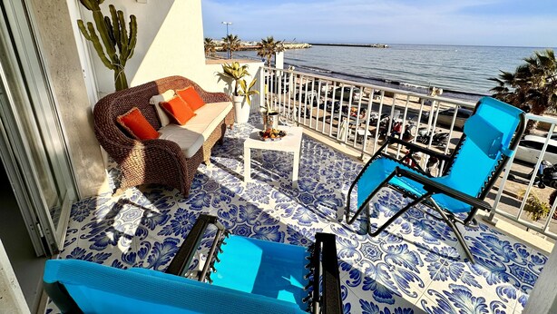 A balcony with a wicker sofa, a blue chair, and a small table with a potted plant. - The terrace by the sea (Sciacca)