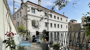 Una terraza en la azotea con plantas en macetas, un banco de metal azul y vistas a un edificio blanco con múltiples ventanas y tejado de tejas rojas.