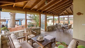 Eine überdachte Terrasse im Freien mit Holzmöbeln, Steinmauern und großen Fenstern mit Blick auf einen Strand.