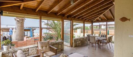 A covered outdoor patio with wooden furniture, stone walls, and large windows overlooking a beach.