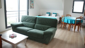 Una habitación con un sofá verde, una cama con ropa de cama azul, una mesa de comedor con sillas y un ventanal grande.