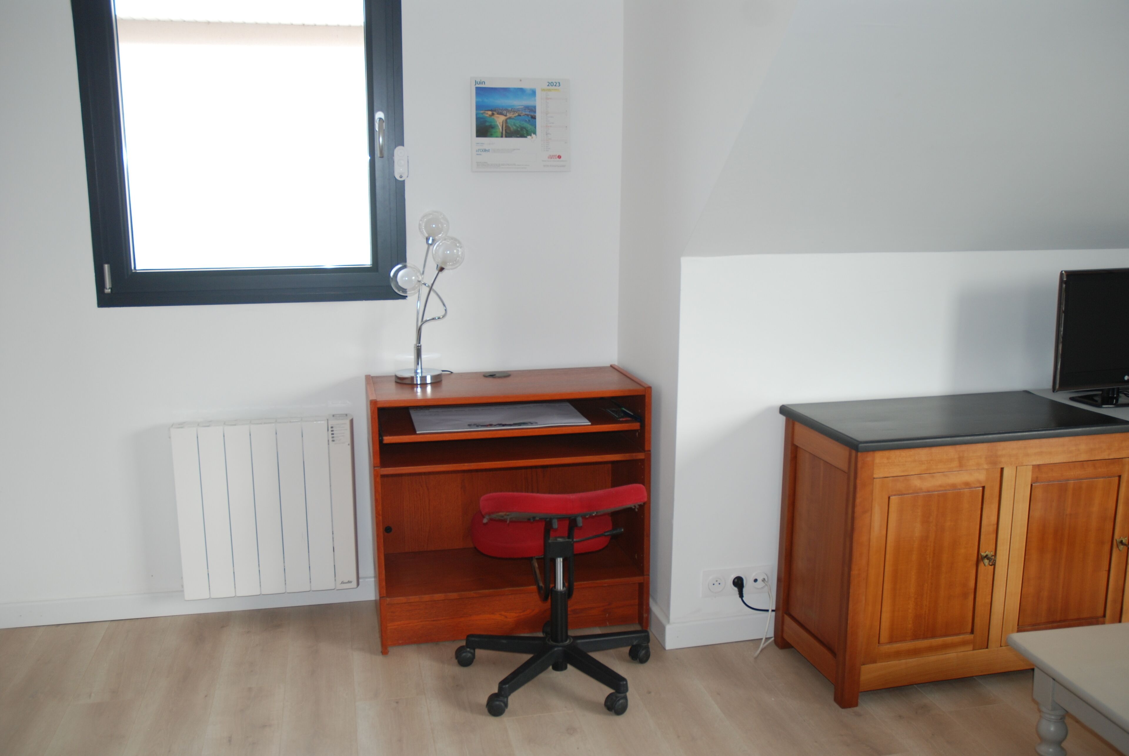 A room with a wooden desk, a red office chair, a radiator, a window, and a cabinet.