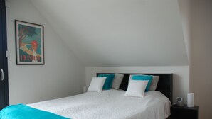 Un dormitorio con cama, almohadas, una mesita de noche, un cuadro enmarcado y una pared blanca.