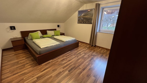 Une chambre avec un plancher en bois, un lit avec une literie verte et blanche, une table de chevet, une lampe fixée au mur et une fenêtre avec des rideaux.
