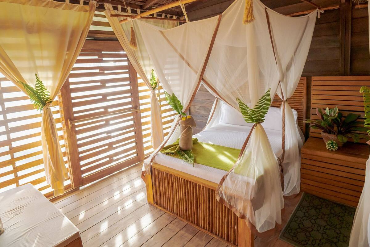 Quarto de madeira com cama sob um dossel, piso de madeira e janela com persianas.